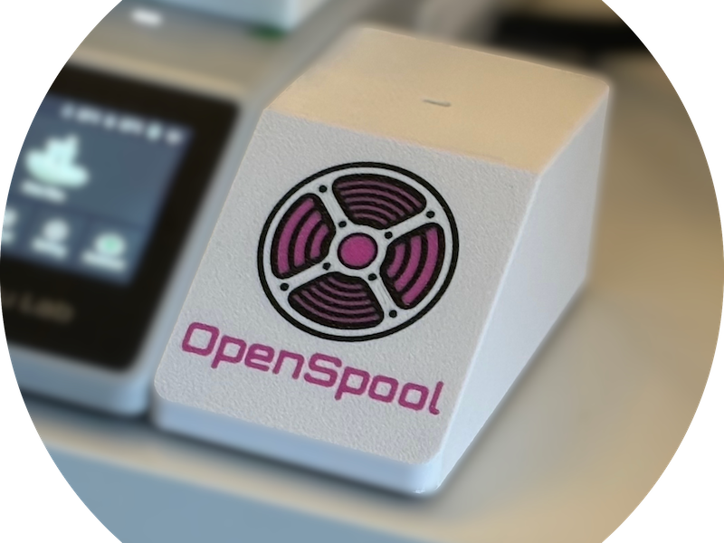 The OpenSpool reader, a small white 3D-printed enclosure with the OpenSpool logo, sitting next to a 3D printer.